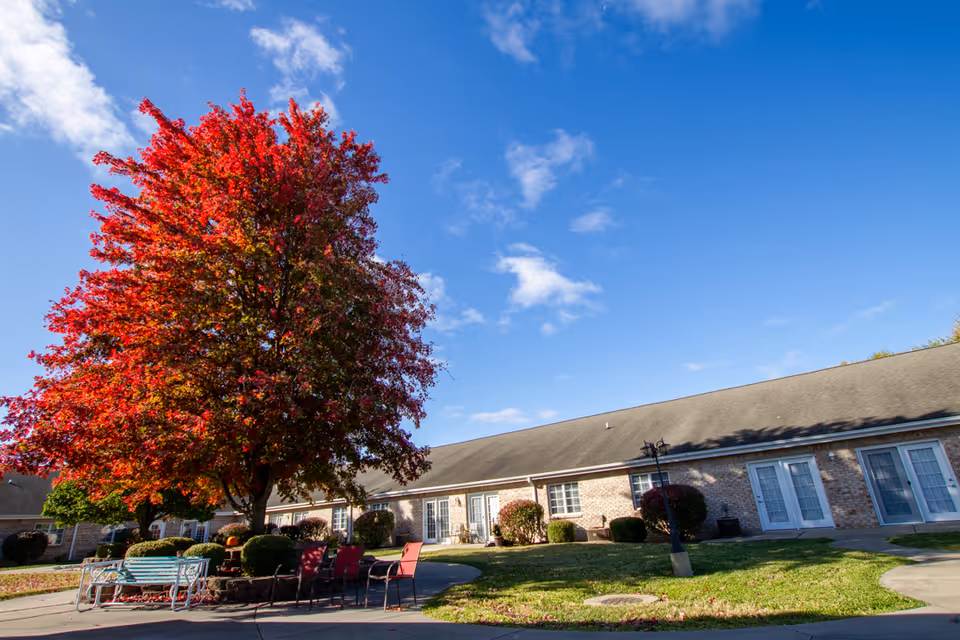 A single-story brick building with multiple doors and windows, surrounded by a well-maintained lawn and bushes. In the foreground, there is a large tree with vibrant red autumn leaves, and outdoor seating including a bench and several chairs arranged around a small table. The sky is clear with a few scattered clouds.