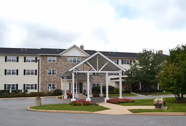 Front entrance of a three-story senior living building with a covered porte-cochere, landscaped flowerbeds, and benches.