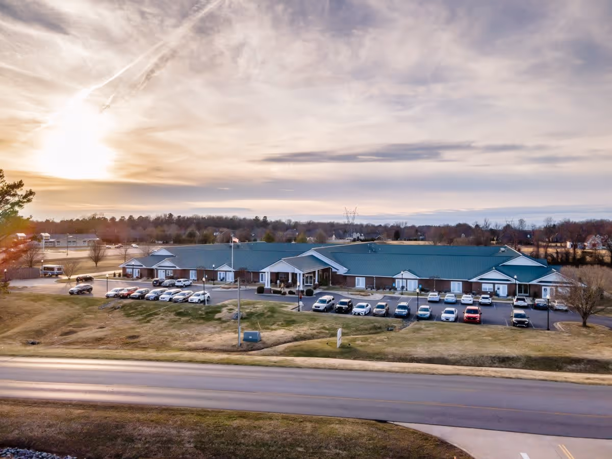 Aerial view of Rivercrest Place, a single-story senior living facility with a green roof and brick exterior, surrounded by a parking lot with several cars and open grassy areas under a partly cloudy sky during sunset.