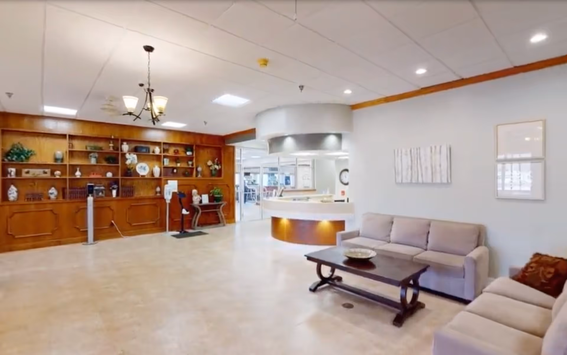 A spacious and well-lit common area in a senior living facility featuring two beige sofas with a dark wooden coffee table in the center. The room has light-colored tiled flooring, a wooden built-in bookshelf filled with decorative items and plants, and a circular reception desk with overhead lighting. The walls are painted light gray with framed artwork, and the ceiling has recessed lighting and a chandelier.