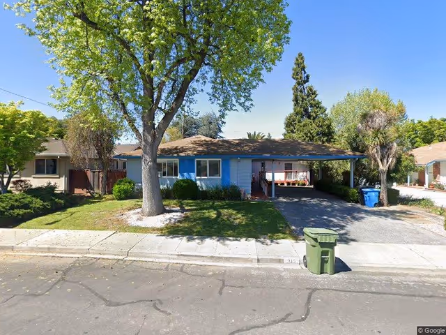 Single-story residential building with a carport on the right side, a large tree in the front yard, green grass, and bushes. The house has blue shutters and a driveway with a green trash bin near the curb. The sky is clear and blue.
