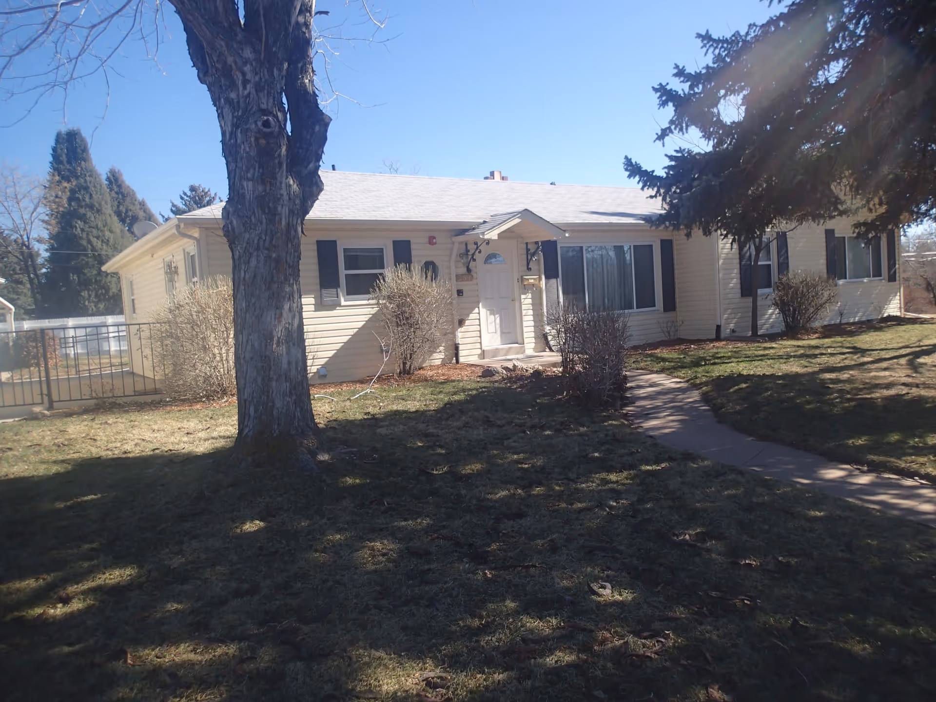 Single-story light-colored house with a front door, windows, large trees, and a walkway across a grassy yard.