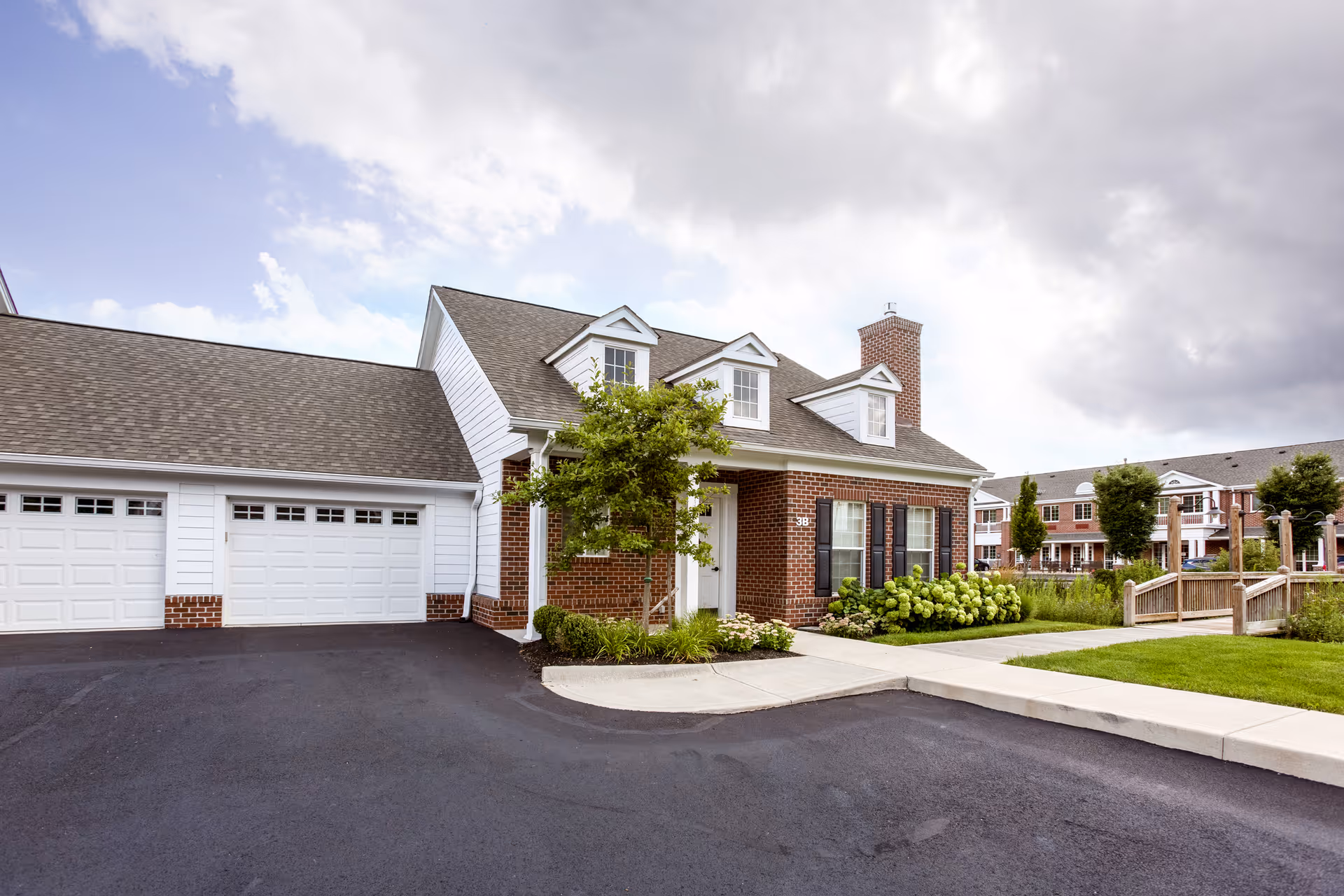 Exterior view of a residential-style building with brick and white siding, featuring a garage with white doors, a small tree, and landscaped greenery in front. The sky is partly cloudy.