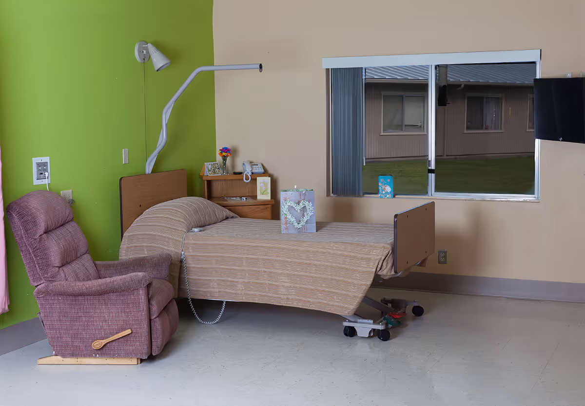 A patient room in a healthcare facility with a hospital bed covered in a beige blanket, a purple recliner chair, a small wooden bedside table with a phone, flowers, and cards, a window with vertical blinds showing an outside view of another building, and a wall-mounted TV.