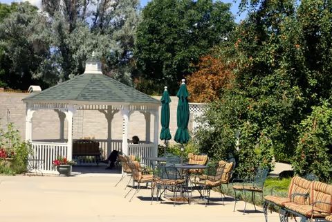 Outdoor patio area with a white gazebo, several metal tables and chairs with cushions, two closed green umbrellas, and surrounding greenery including trees and bushes.