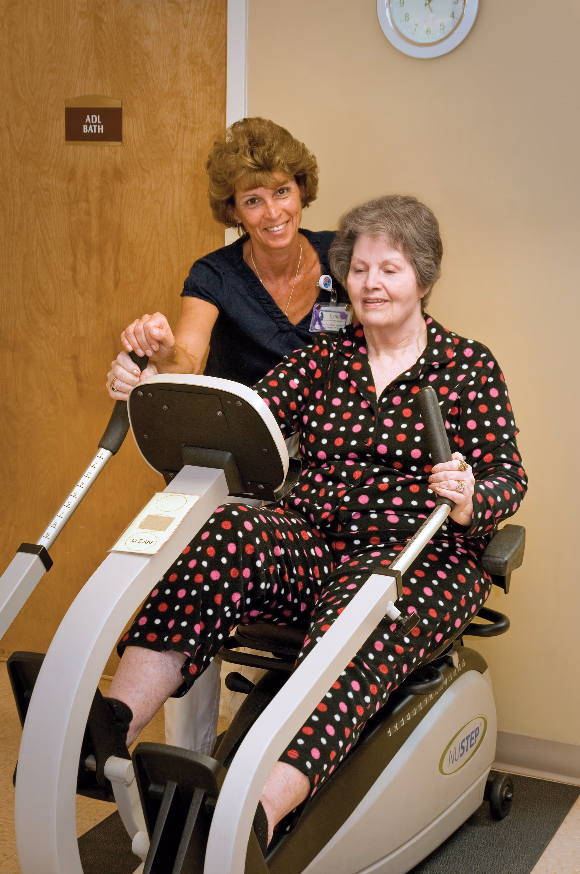 An elderly woman in a black outfit with pink and white polka dots is seated on a NuStep exercise machine, using the handles and pedals. A smiling caregiver or physical therapist stands behind her, providing support and encouragement. They are indoors near a door labeled 'ADL BATH' and a wall clock is visible above them.