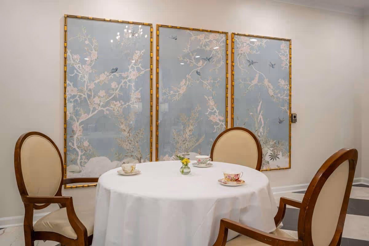 A round table covered with a white tablecloth set with three teacups and saucers, and a small vase with yellow flowers in the center. Three wooden chairs with beige cushioned seats and backs surround the table. Behind the table, there are three large decorative panels with floral and bird designs in soft pastel colors framed in gold, mounted on a light-colored wall.