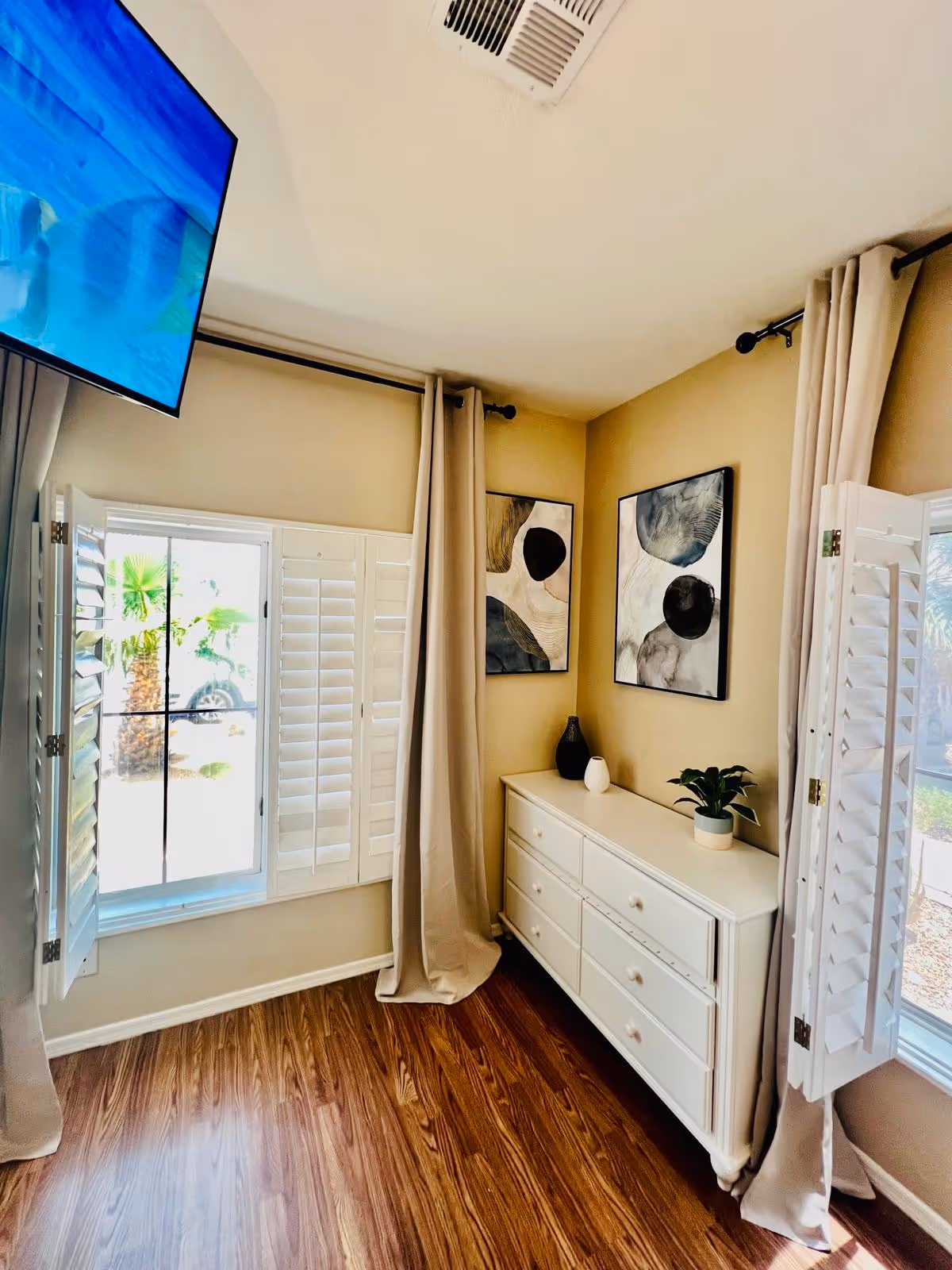A corner of a room with light beige walls and wood flooring. There is a white dresser with six drawers, decorated with a small plant and two vases. Above the dresser are two abstract art paintings. The room has two windows with white plantation shutters and beige curtains. A ceiling vent and a wall-mounted TV are also visible.