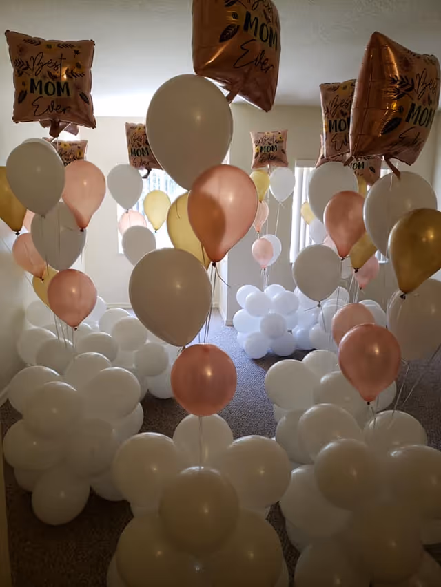 A carpeted interior room filled with clusters of white, pink, and gold helium balloons, including rose-gold 'Best Mom Ever' balloons.