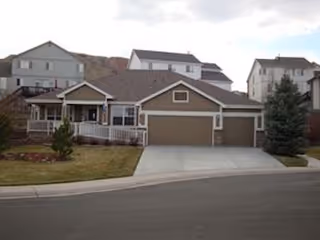 A single-story residential house with a three-car garage, a front porch, and a small lawn with a few trees. Several two-story houses are visible in the background under a partly cloudy sky.