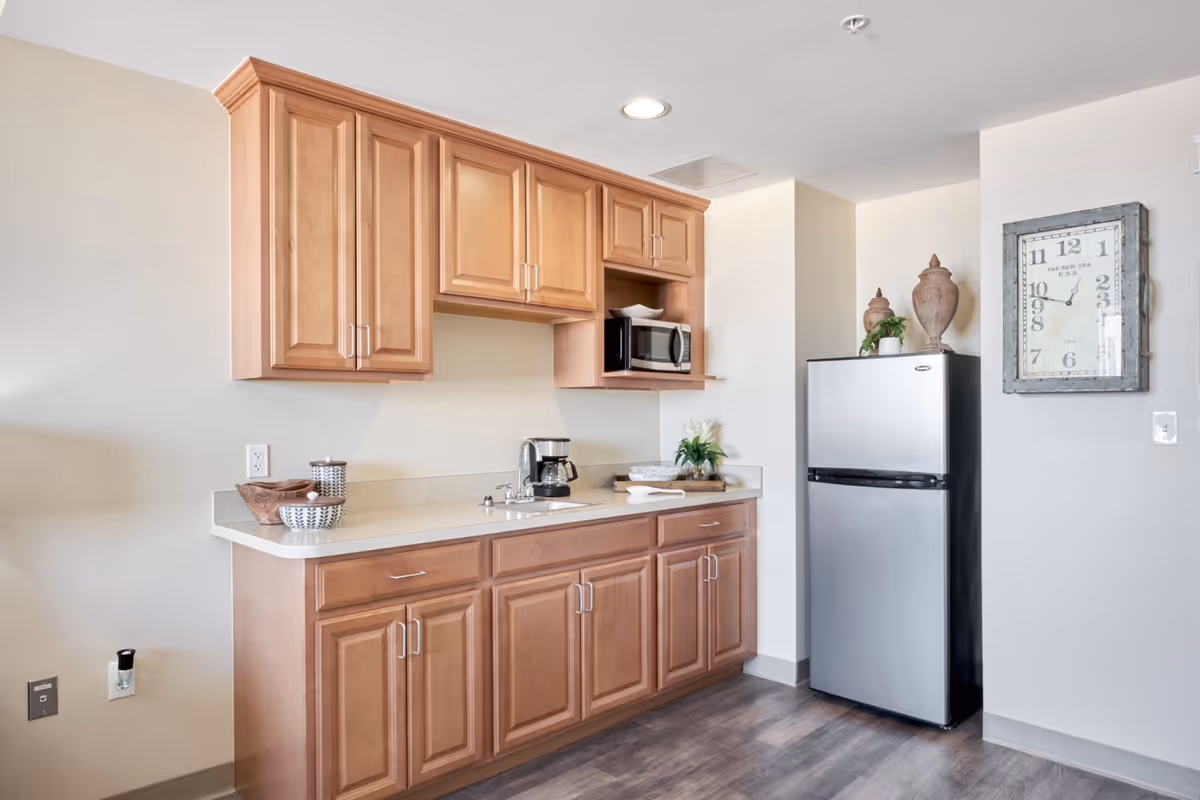 A small kitchen area with light wood cabinets, a countertop with a coffee maker and decorative bowls, a stainless steel refrigerator, and a wall clock. The floor is dark wood, and the walls are light-colored.