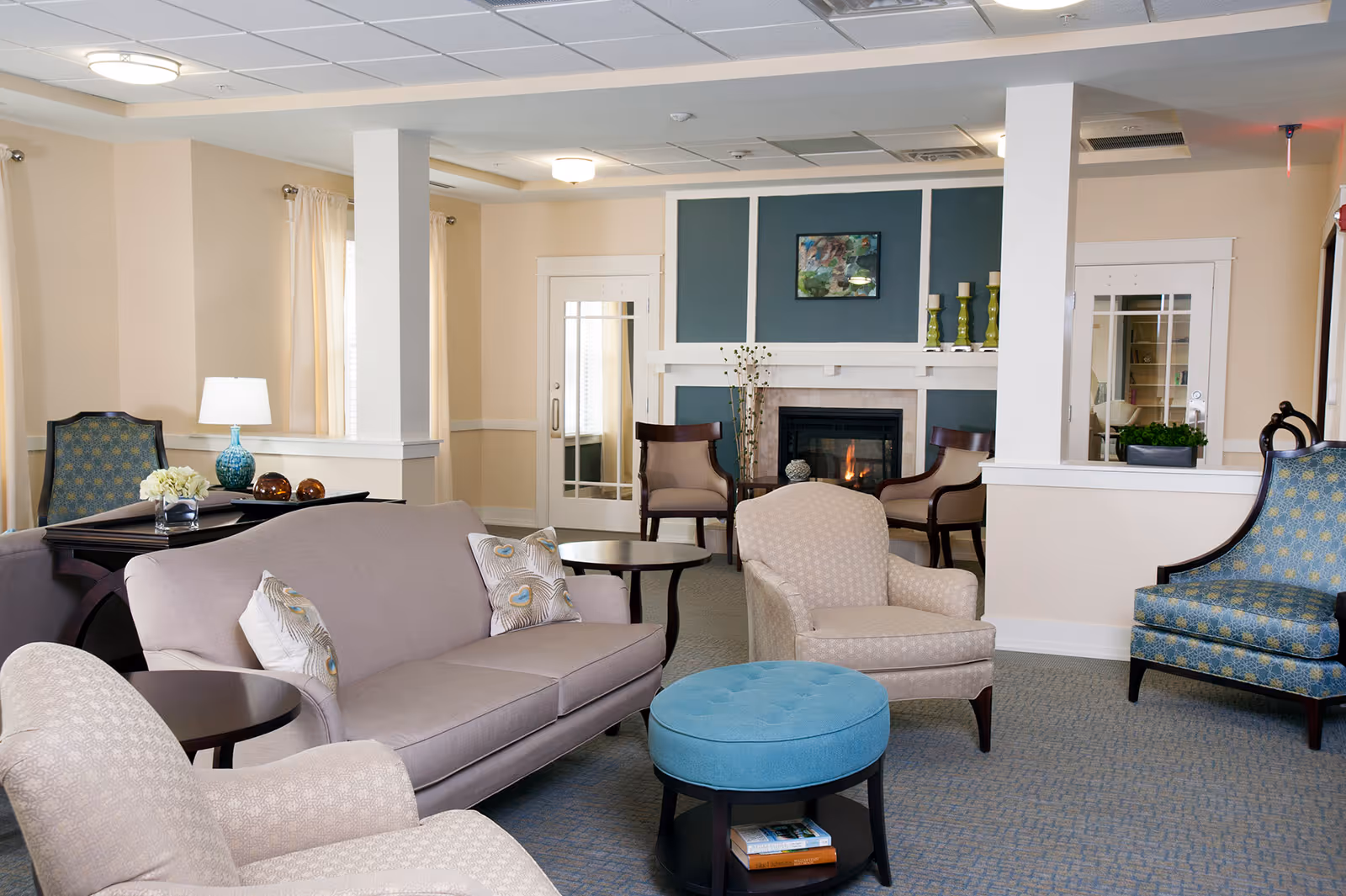 A cozy living room area in a senior living facility featuring a beige sofa with decorative pillows, two beige armchairs, a blue patterned armchair, and a round blue ottoman with a shelf underneath holding books. The room has a fireplace with a fire burning, decorated with green candle holders and a framed artwork above it. There are two white pillars, cream-colored walls, and windows with light curtains allowing natural light to enter.