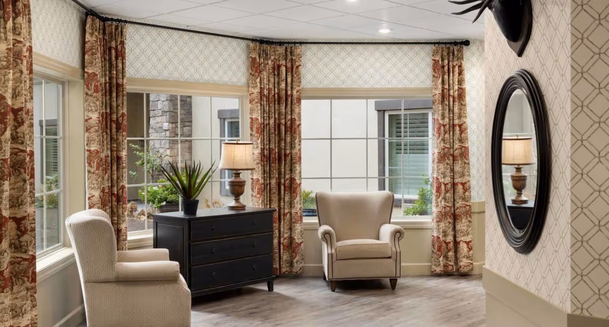 A cozy sitting area in a senior living community with two beige upholstered armchairs, a black chest of drawers with a potted plant and a table lamp on top, large windows with patterned curtains, and a round mirror on the wall reflecting the lamp.