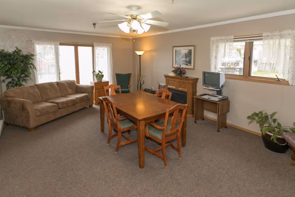 A bright common room with a wooden dining table and chairs, a sofa, TV on a small desk, a fireplace, plants, and a ceiling fan.