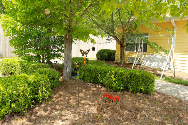 Landscaped yard with hedges, trees, a butterfly garden stake, red flowers, and a porch swing beside a yellow building.