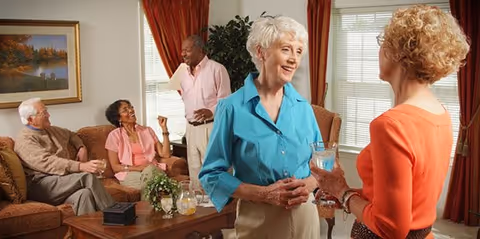A group of five elderly people socializing in a warmly decorated living room. Two women are standing and talking near a window with red curtains, one holding a glass of water. Three others are seated on couches around a wooden coffee table with a plant and a pitcher of water. The room has a framed landscape painting on the wall.