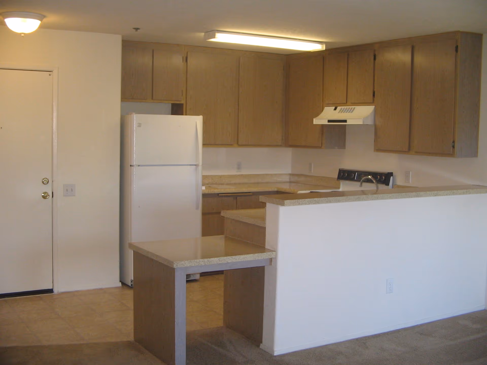 Interior view of a kitchen area in a senior living facility named Villa Pacifica. The kitchen features light wood cabinets, a white refrigerator, a stove with a range hood, and a countertop with an extended bar area. The floor is tiled near the kitchen and carpeted in the adjacent area. A closed door is visible on the left side of the image.