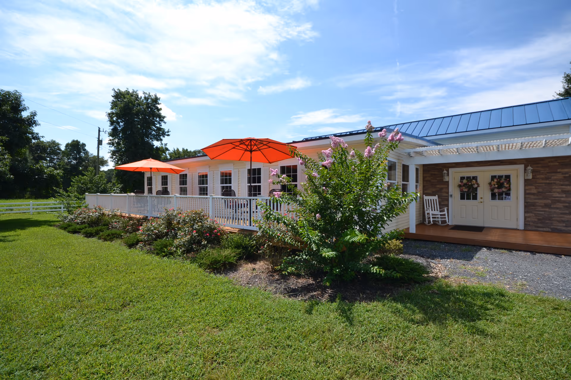 Exterior view of Chesapeake Cottage Assisted Living showing a single-story building with a blue metal roof, white siding, and a wooden porch. The porch has white railings, two orange umbrellas, and several chairs. There are flowering bushes and green grass in the foreground under a partly cloudy sky.