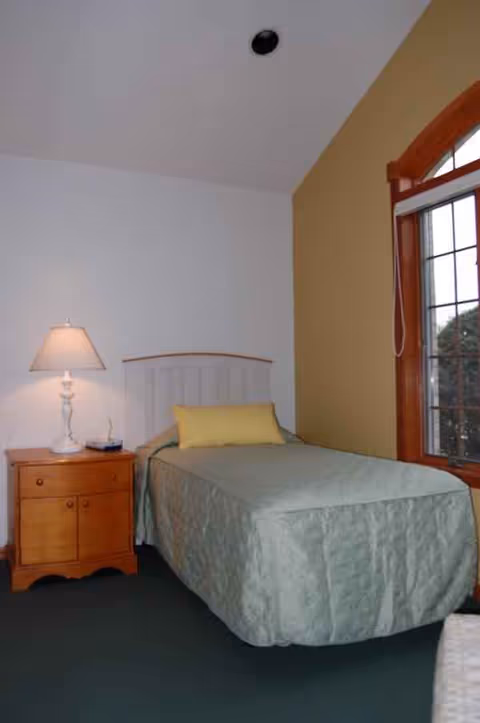 Single bed with quilt, wooden nightstand, lamp, and window in a bedroom.