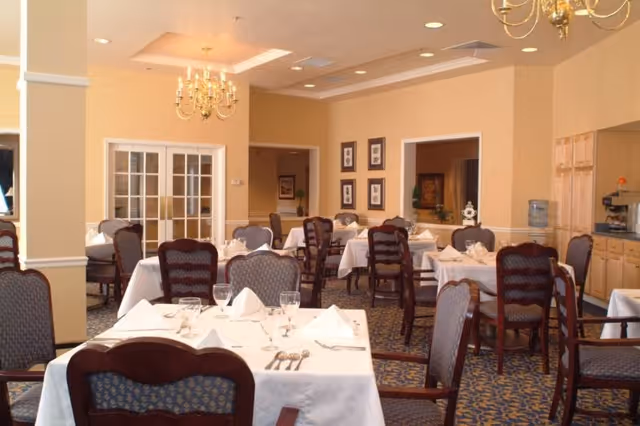 Formal dining room with tables covered in white tablecloths, set with glassware and silverware beneath chandeliers.