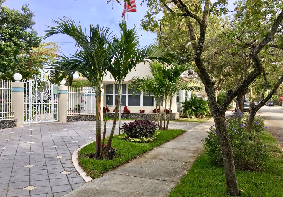 Exterior view of Azalea Gardens Assisted Living Facility showing a paved driveway, a white decorative gate with the initials AG, palm trees, green grass, and a sidewalk lined with trees and flowering plants under a partly cloudy sky.