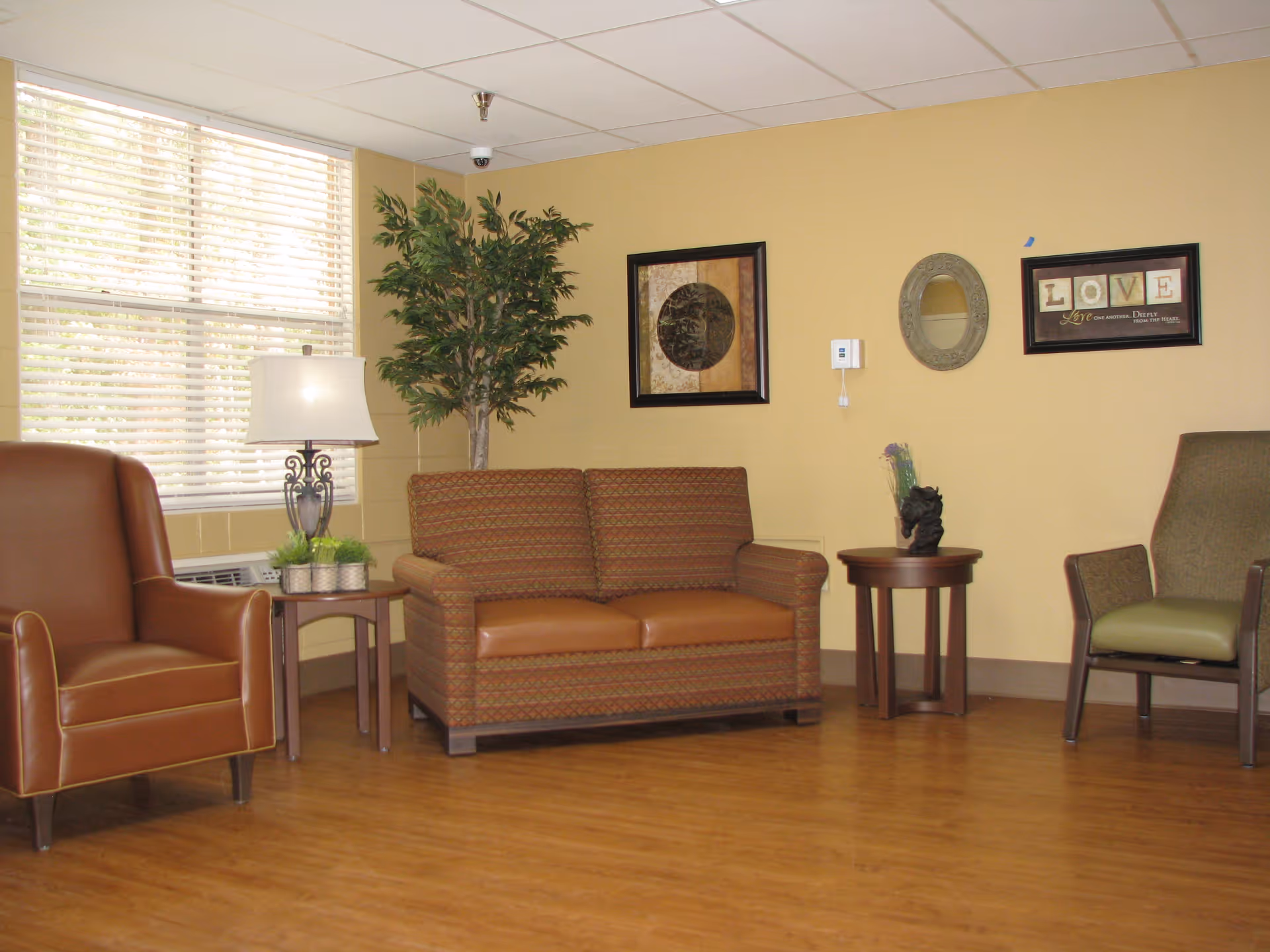 A cozy living room area with a brown leather armchair, a patterned loveseat, and a green cushioned chair. There are two small wooden side tables, one with a lamp and small plants, and the other with a decorative item. A large window with blinds lets in natural light. The walls are painted beige and decorated with framed artwork and a round mirror.