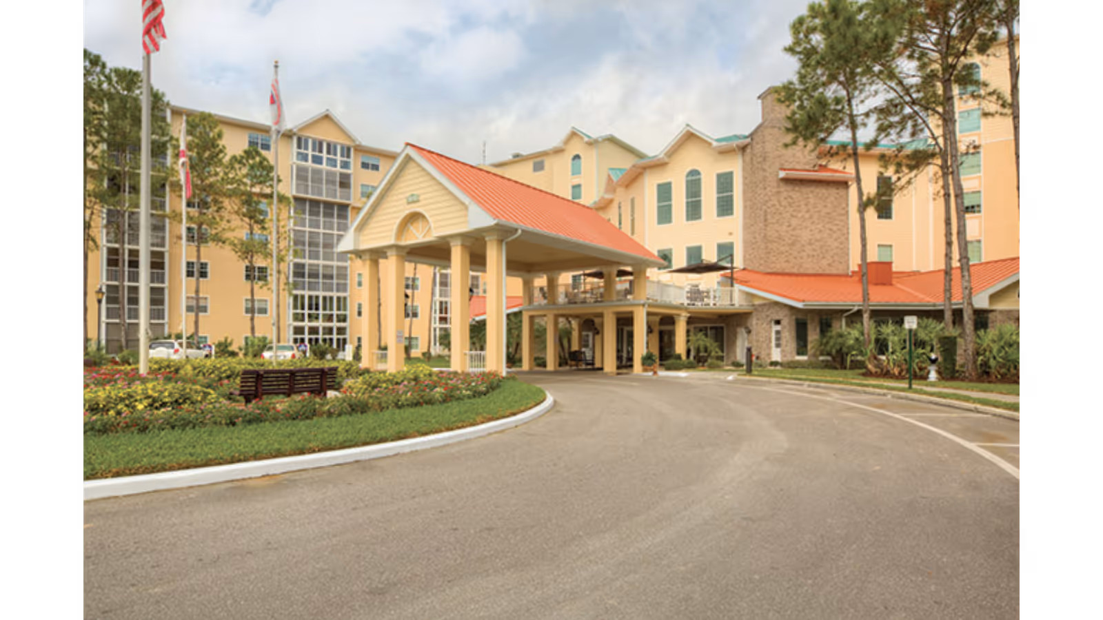 Exterior view of a senior living facility named Freedom Plaza featuring a multi-story building with beige and brick walls, green window shutters, and a red roof. The entrance has a covered drop-off area supported by columns. There are landscaped gardens with flowers and trees, a curved driveway, and flagpoles with flags near the entrance.
