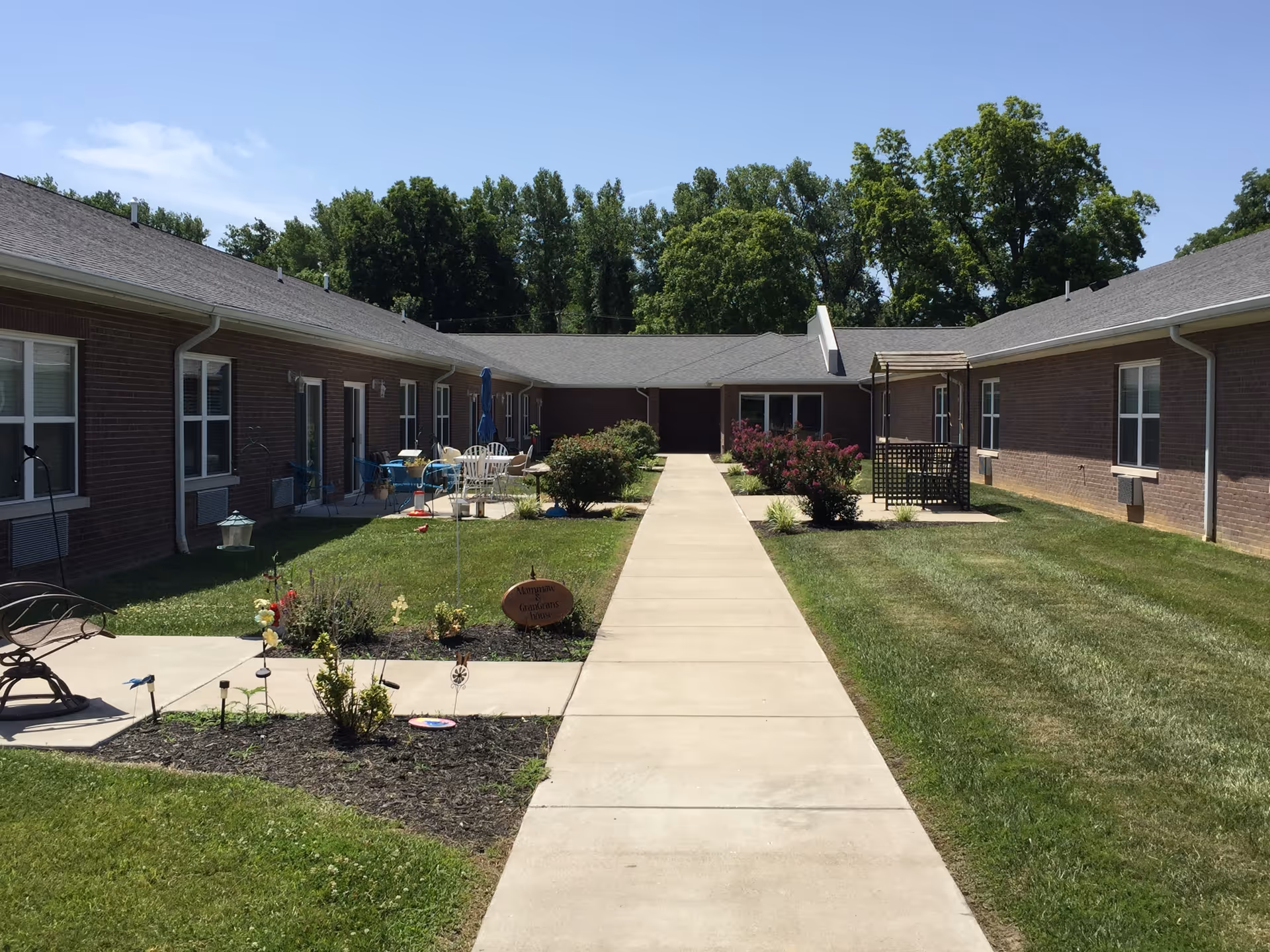 Sunlit courtyard with a central concrete walkway flanked by grass, flowerbeds, patio seating, and single-story brick residential wings.