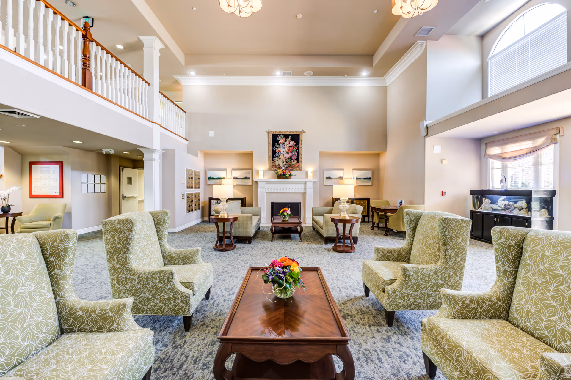 A spacious and well-lit living room area in a senior living facility with high ceilings and large windows. The room features multiple green patterned armchairs arranged around wooden coffee tables with floral centerpieces. There are two sofas facing a white fireplace adorned with a floral tapestry and a flower arrangement. The room also includes side tables with lamps, framed artwork on the walls, and an aquarium near a window with blinds.