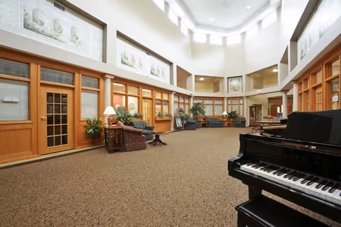 Spacious senior living facility common area with high ceilings and large windows near the ceiling. The room features a patterned carpet, wooden framed glass doors and windows, several seating areas with armchairs and plants, and a black grand piano in the foreground.