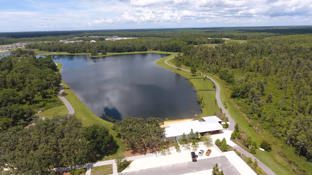 Aerial view of a large pond surrounded by green grass and trees with a paved walking path curving around the pond. There is a small building with a white roof near the pond and a parking area with a few cars. The surrounding area is heavily wooded under a partly cloudy sky.