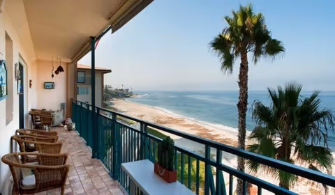 Balcony with wicker chairs and a small table overlooking a sandy beach and ocean, with palm trees visible near the shore under a clear sky.