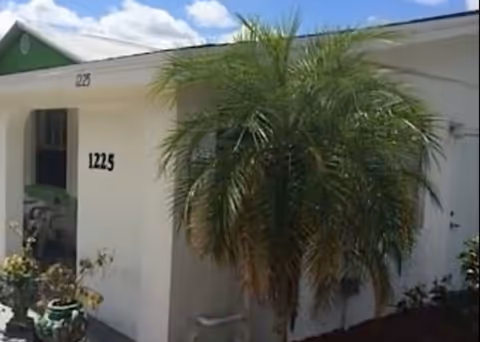 Exterior view of a single-story white building with the number 1225 displayed near the entrance. A palm tree and some potted plants are visible near the doorway under a partly cloudy sky.
