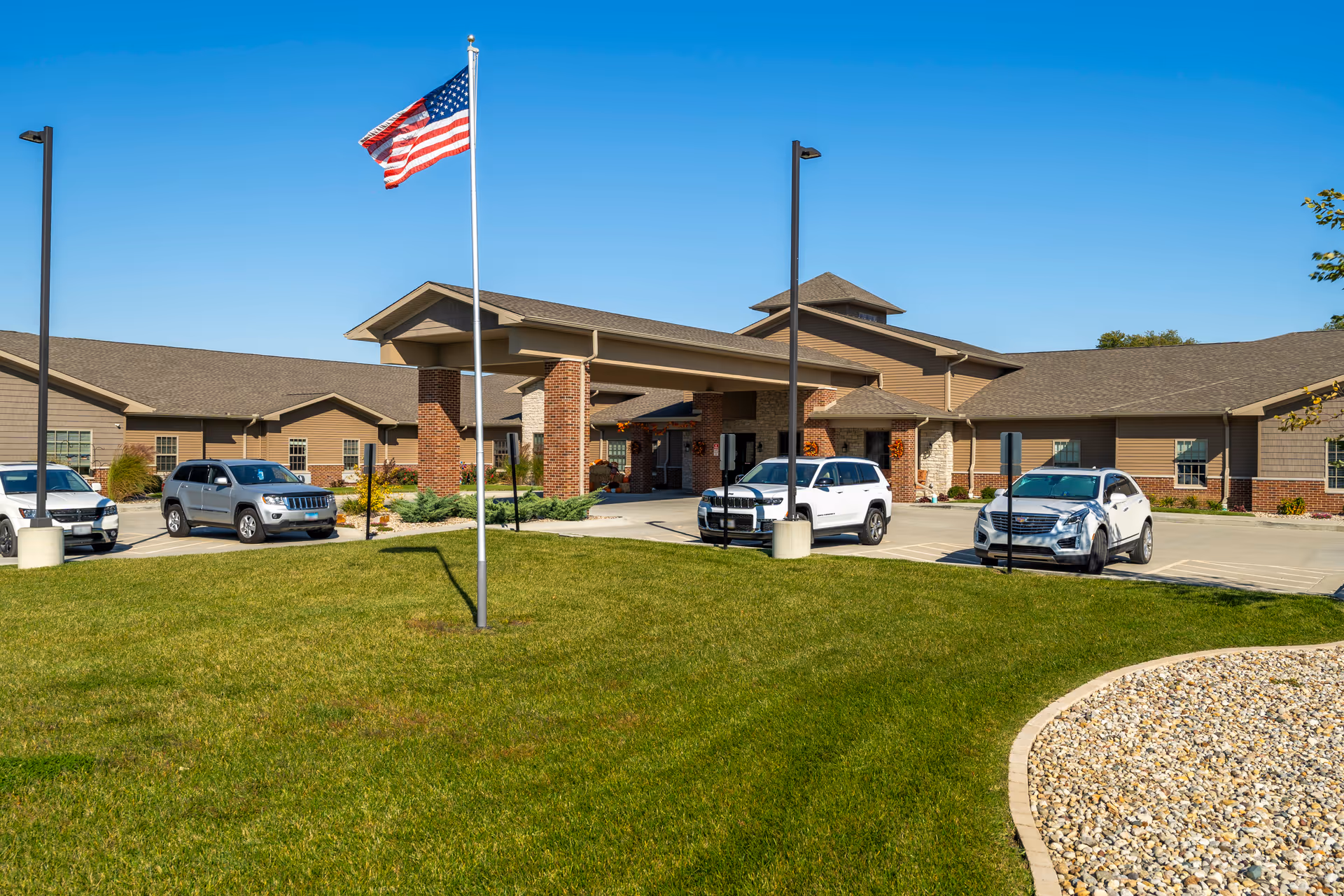 Front exterior of a single-story senior living facility with an American flag on a lawn and several parked cars near the entrance.