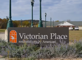 Entrance sign reading "Victorian Place" with lamp posts, landscaping, and the facility building and parked cars in the background.