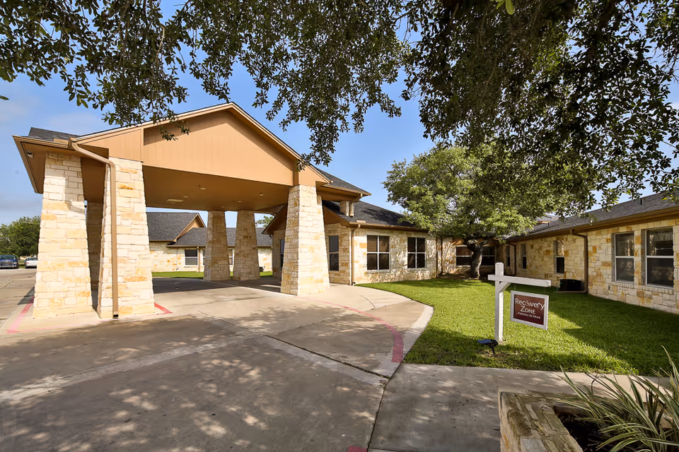 Exterior view of Cuero Nursing and Rehabilitation Center showing a covered entrance with stone pillars, a driveway, green lawn, trees, and a sign that reads 'Recovery Zone Patients at Work'.