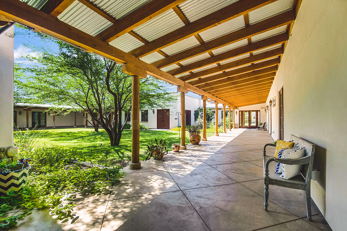 Covered outdoor walkway with wooden beams and a corrugated roof, overlooking a green courtyard with trees and plants. A bench with decorative pillows is placed along the wall on the right side.