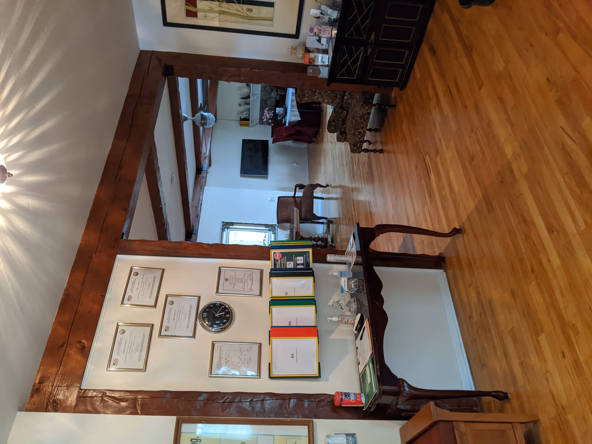 Interior view of a senior living facility reception or common area with wooden floors and exposed wooden beams. A wall features framed certificates and a clock above a table holding colorful binders, hand sanitizer, and other small items. In the background, there are chairs, a TV mounted on the wall, and a cabinet with various items on top.