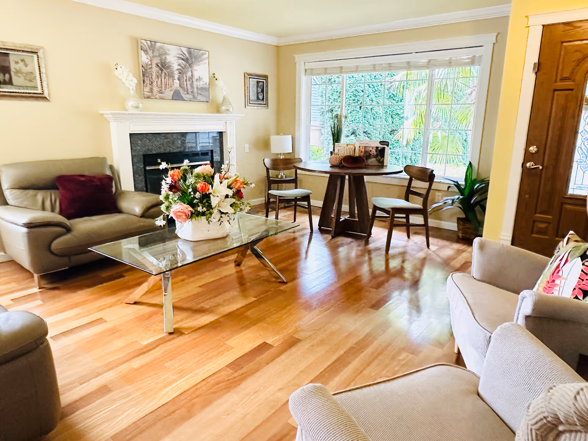 A bright and cozy living room with wooden flooring, a glass coffee table with a floral arrangement, beige leather and fabric armchairs, a fireplace with decorative vases and framed artwork above it, a round wooden table with two chairs near a large window showing greenery outside, and a wooden front door with decorative glass.