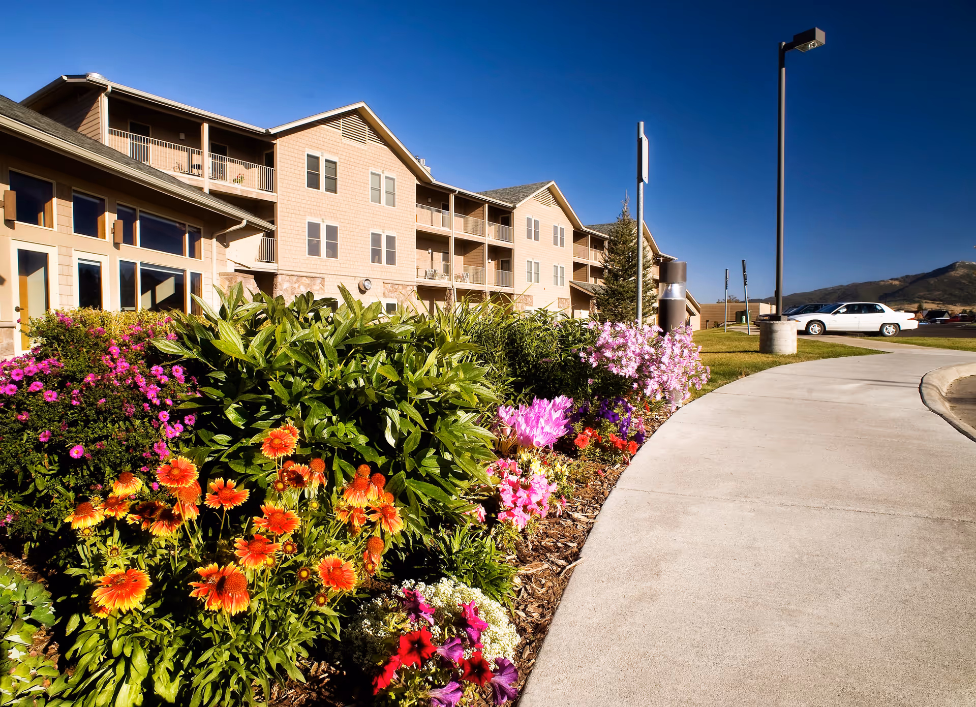 Exterior view of Bozeman Health Hillcrest Senior Living, Aspen Pointe building with a clear blue sky, a curved concrete pathway, and a colorful flower bed with various blooming flowers in the foreground. A white car is parked in the distance near the building.