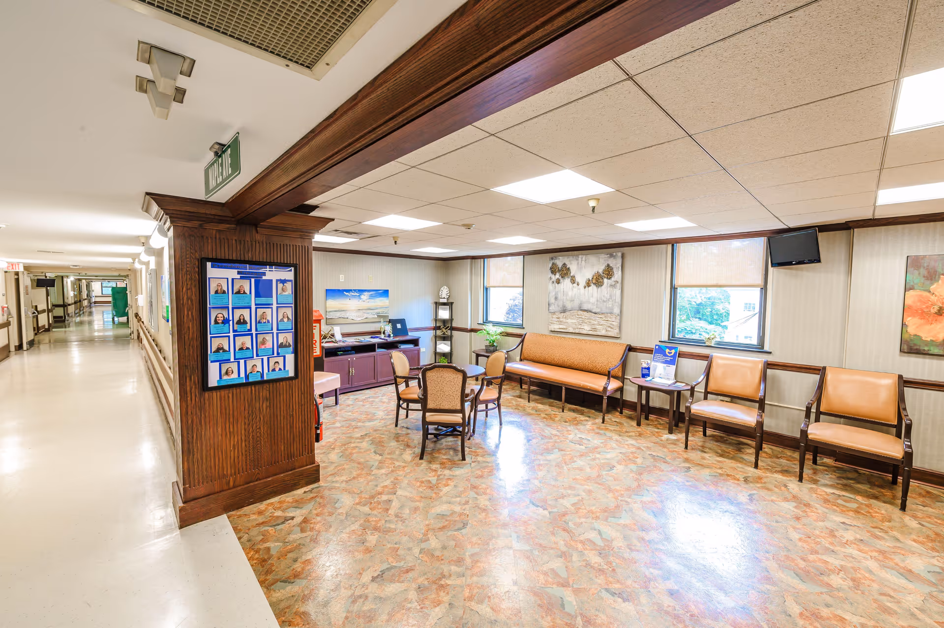 A bright waiting area inside a senior living facility with several chairs and a small table arranged near two windows. The walls are decorated with paintings and a bulletin board with staff photos is mounted on a wooden column. The floor has a patterned linoleum surface and the ceiling has recessed lighting. A hallway extends to the left with handrails along the walls.