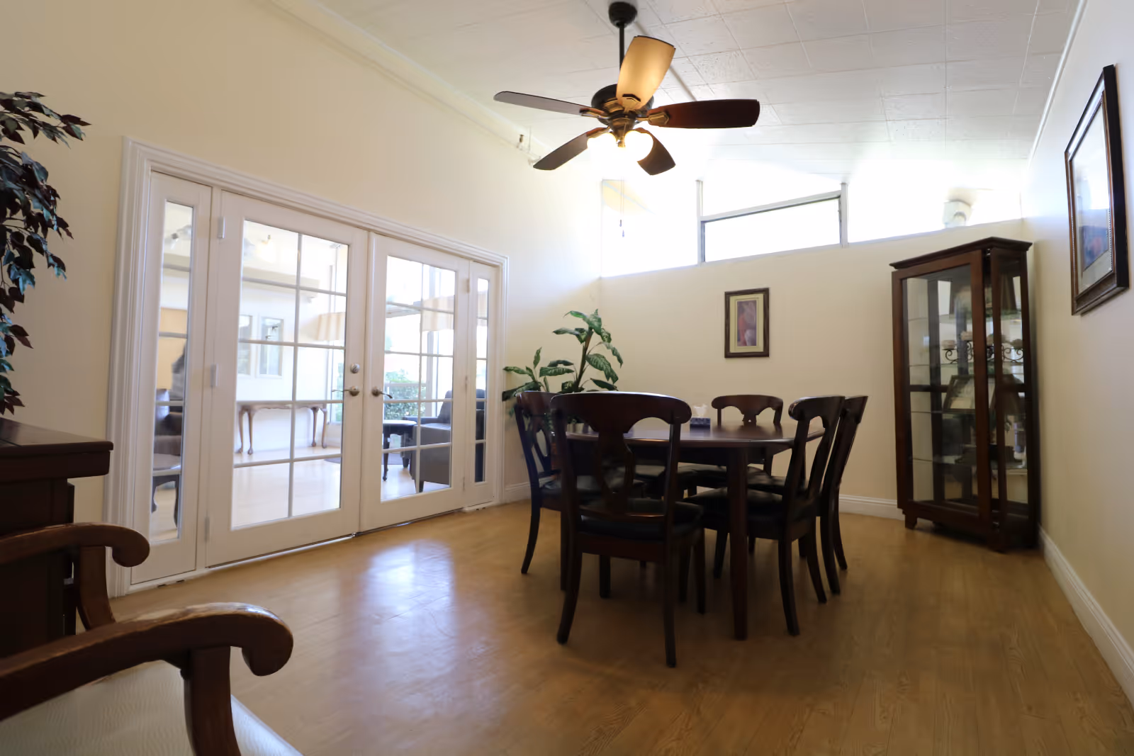 A bright dining area with a round wooden table and chairs, a glass-front cabinet, ceiling fan, and French doors to an adjoining room.