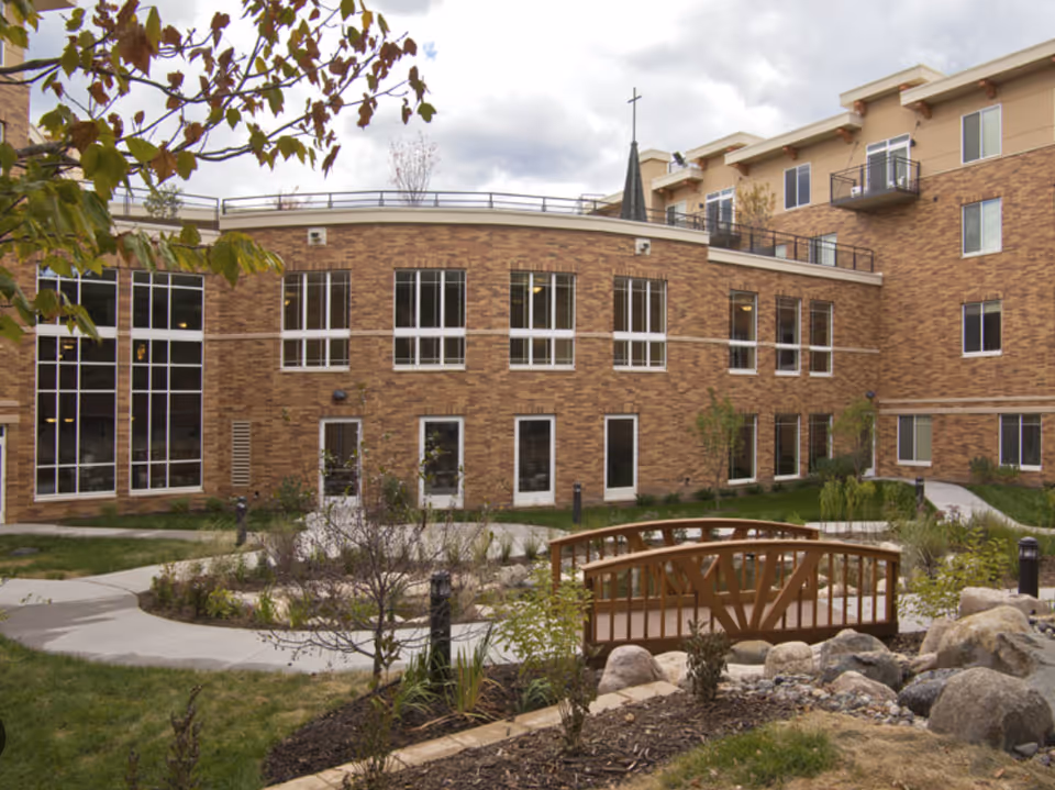 Outdoor courtyard area of Carondelet Village featuring a small wooden footbridge over a dry rock bed, surrounded by landscaped plants and a curved concrete walkway. The background shows a multi-story brick building with large windows and balconies under a cloudy sky.