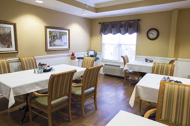 A dining room with several tables covered with white tablecloths and surrounded by wooden chairs with green and beige striped cushions. The room has light yellow walls, two framed paintings, a window with a valance, a wall clock, and a wooden floor.