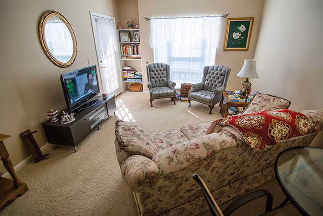 A cozy living room with a floral patterned sofa adorned with red and gold pillows, two gray tufted armchairs near a window with sheer white curtains, a wooden TV stand with a flat-screen television, a round glass-top table with chairs, a wall-mounted oval mirror, and a small bookshelf in the corner.
