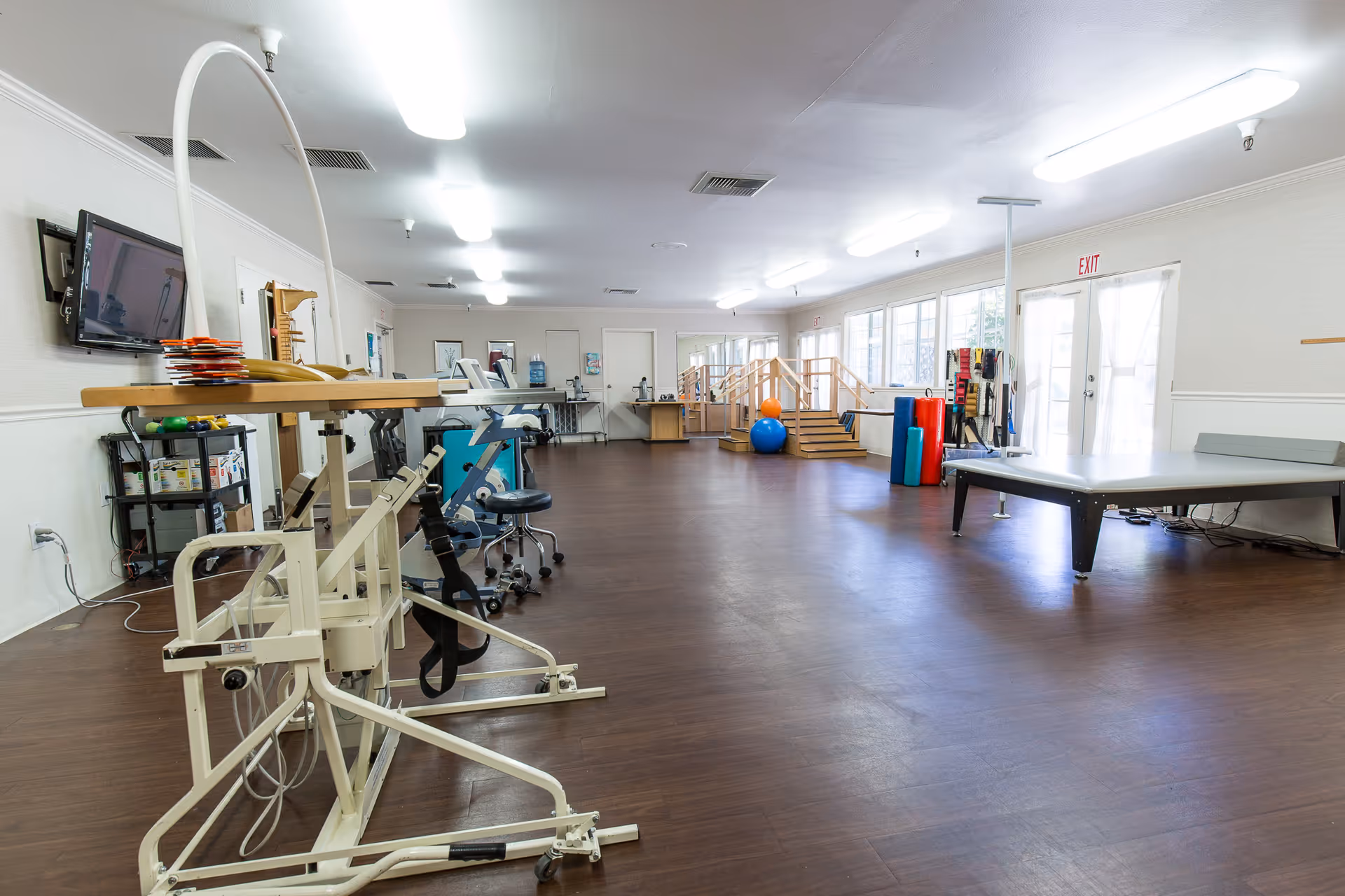 A spacious rehabilitation or physical therapy room with various exercise and therapy equipment, including a parallel bar setup, therapy balls, foam rollers, and a table. The room has large windows letting in natural light, wooden flooring, and bright overhead lighting.