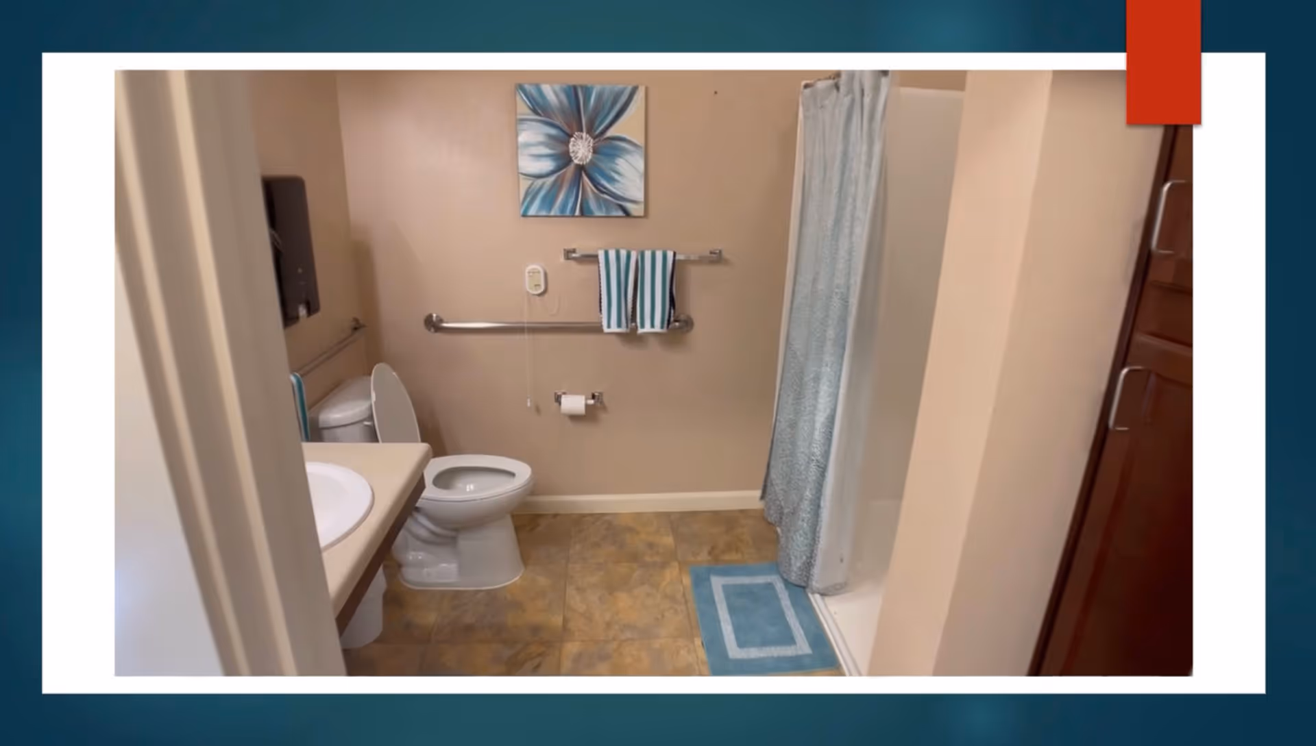 A bathroom with a toilet, sink, and shower area. The toilet has a grab bar beside it, and there is a towel rack with two striped towels above the toilet paper holder. A blue and white floral painting hangs on the wall above the towel rack. The shower has a light blue curtain and a matching blue bath mat on the floor. The floor is tiled in a beige pattern.