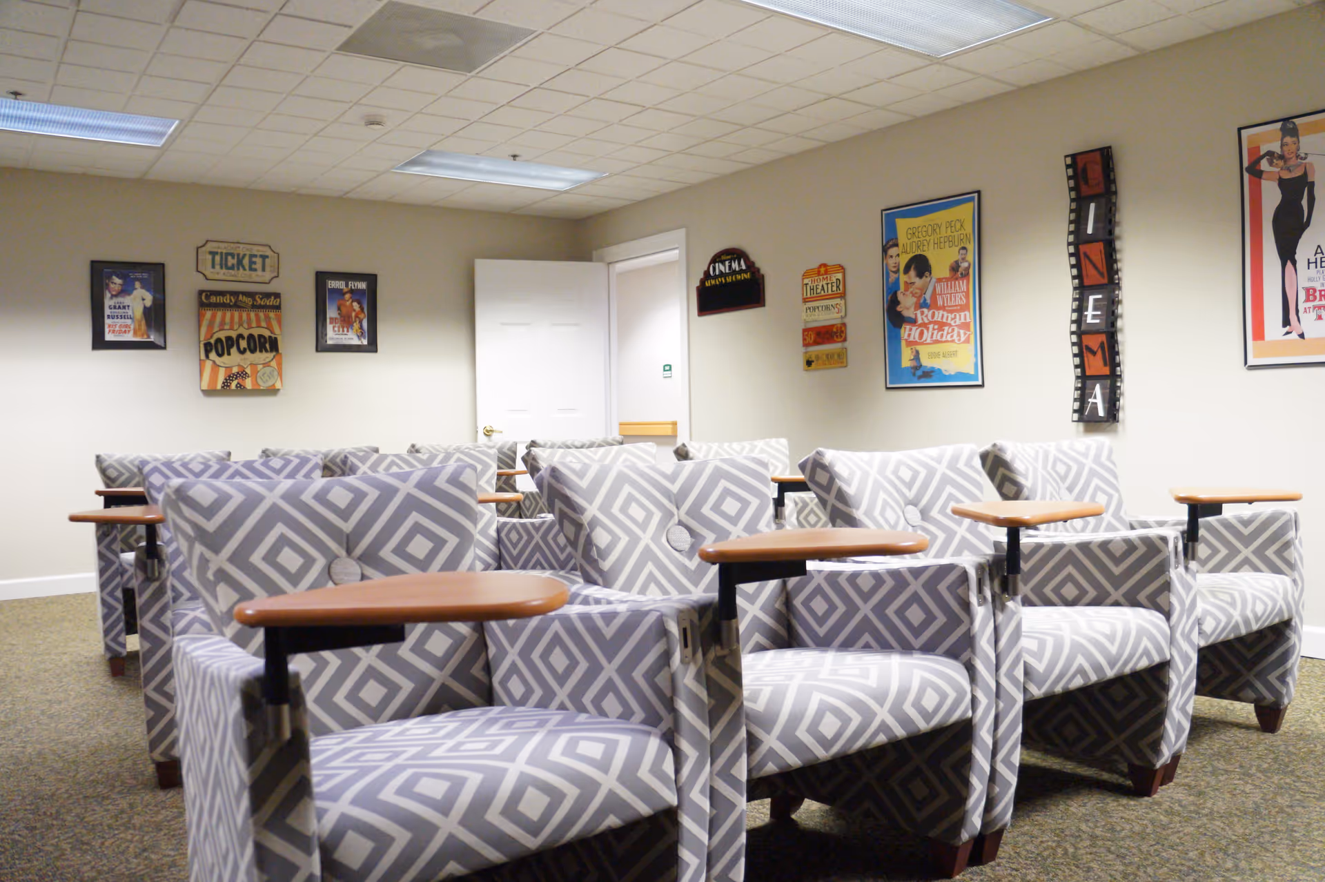 A small theater room with multiple patterned armchairs arranged in rows facing a blank wall. The walls are decorated with vintage movie posters and cinema-themed signs. The room has a carpeted floor and a white door in the background.