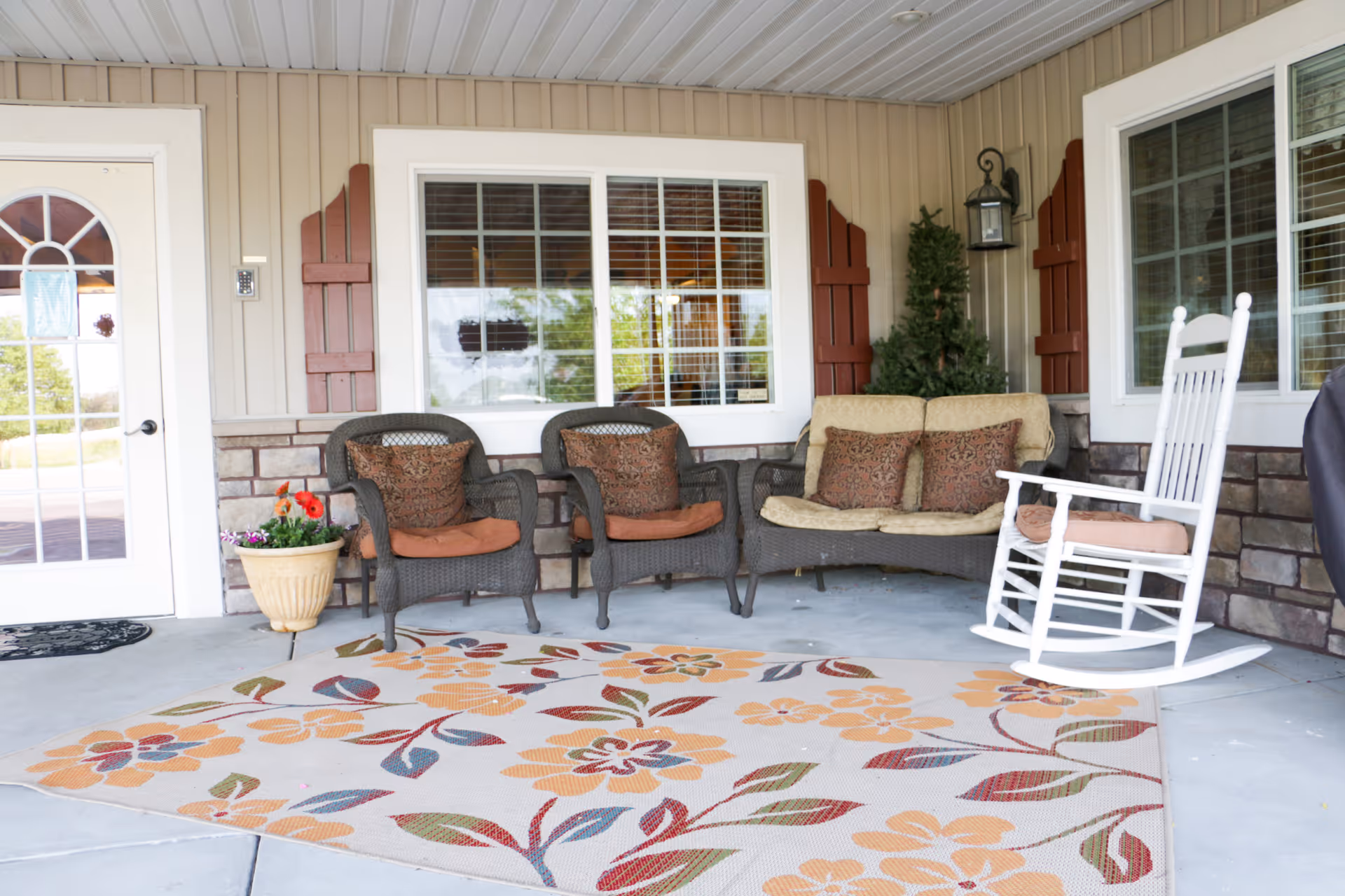 A covered outdoor patio area with a floral patterned rug, two wicker armchairs with cushions, a wicker loveseat with cushions, a white wooden rocking chair, a potted plant with flowers, and a small artificial tree in the corner. The patio has beige siding, stone accents, and windows with white frames and red shutters.