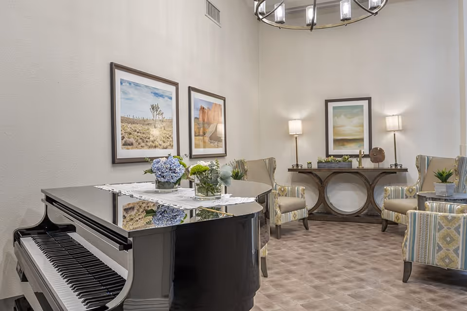 A cozy living room area featuring a black grand piano with floral arrangements on top, several upholstered armchairs with patterned fabric, a wooden console table with decorative items, framed landscape artwork on the walls, two standing lamps, and a modern chandelier hanging from the ceiling.